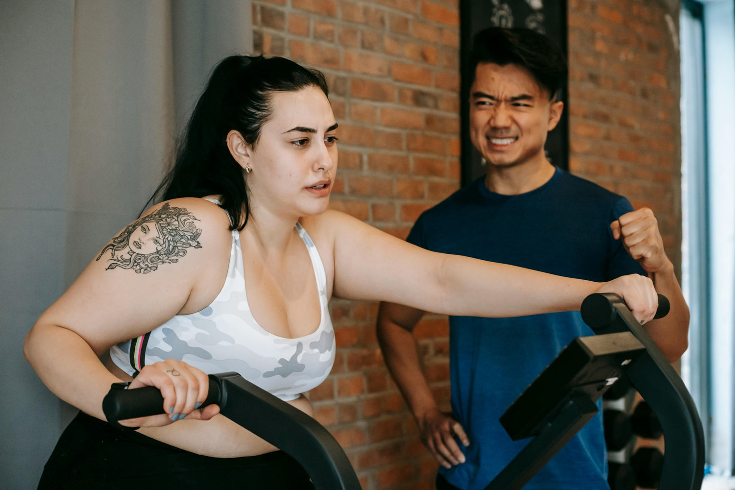 Woman struggles to pedal elliptical machine while trainer watches.